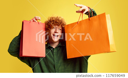 Portrait of young man holding shopping packages, expressing happiness and delight against yellow background. Concept of Black Friday 117659049