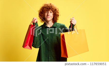 Portrait of delight man, student with curly, red hair, holding packages with gifts against yellow background. Concept of Black Friday Portrait of delight man, student with curly, red hair, holding packages with gifts against yellow background. Concept of Black Friday 117659051