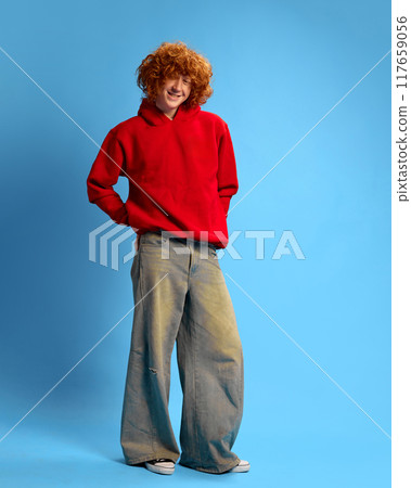 Smiling with satisfaction, young man with red curly hair, shopper dressed in bold red hoodie against blue studio background. Concept of Black Friday sale. 117659056