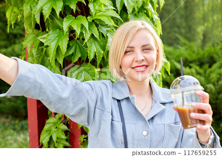 Stylish young blonde walks around the city, drinks a cold drink and takes a selfie on her mobile phone. 117659255