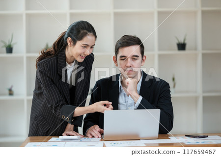 A woman and a man are sitting at a desk looking at a laptop 117659467