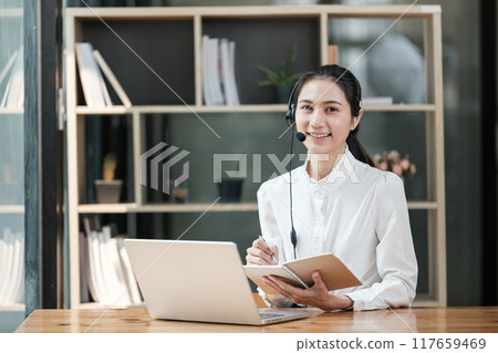 A woman is sitting at a desk with a laptop and a notebook 117659469