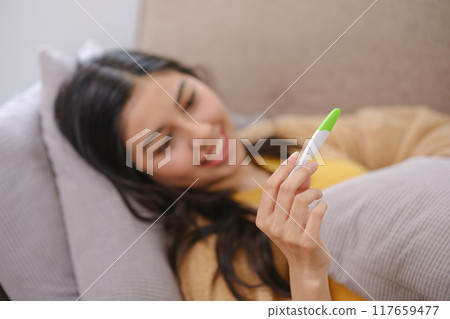 A woman is laying on a couch with a thermometer in her hand A woman is laying on a couch with a thermometer in her hand 117659477