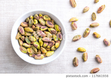 Pistachios kernels, roasted and salted nuts in a white bowl on linen fabric. Shelled fruits of Pistacia vera, crunchy toasted and salted. Ready-to-eat snack food. Close-up, from above, food photo. 117661938