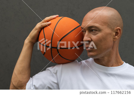 Portrait of handsome sporty adult man in white t-shirt holding basketball orange ball and looking at him near the dark wall. Focus on ball. Portrait of handsome sporty adult man in white t-shirt holding basketball orange ball and looking at him near the dark wall. Focus on ball. 117662156
