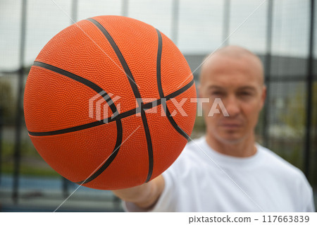 Portrait of sporty adult man holding orange basketball ball in hand on sports court. Focus on ball. Close up. 117663839