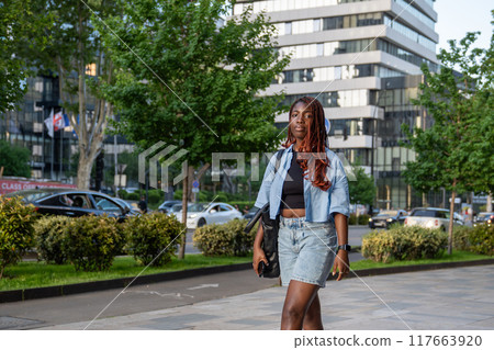 Peaceful black african woman walking along sidewalk with modern downtown buildings in background. 117663920