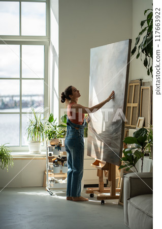 Peaceful woman artist in rays of bright light warm sun moving easel with large canvas to side. Peaceful woman artist in rays of bright light warm sun moving easel with large canvas to side. 117663922
