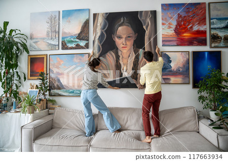 Painters couple at home studio hang artwork on wall. Guy girl together make picture composition Painters couple at home studio hang artwork on wall. Guy girl together make picture composition 117663934