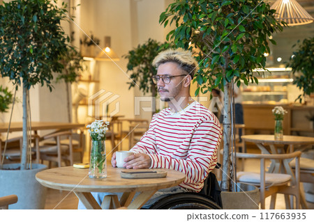 Dreamy reflective disabled man in wheelchair sits with coffee at cafe table. Waits for friend alone 117663935