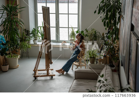 Creative happy female artist in home art studio filled with green flowers and plants in pots. 117663936