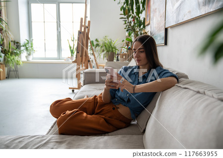 Peaceful pleased woman using smartphone, procrastinating relaxing resting on couch at home workshop Peaceful pleased woman using smartphone, procrastinating relaxing resting on couch at home workshop 117663955