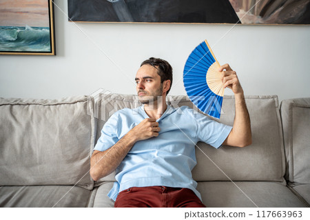 Sad man suffering from heat in apartment, fanning himself with paper fan, no air conditioner at home 117663963