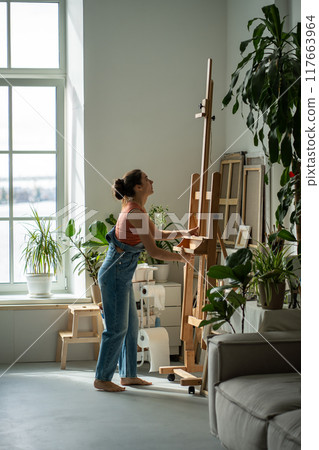 Inspired woman artist preparing easel before work painting process in sunny home studio with plants 117663964
