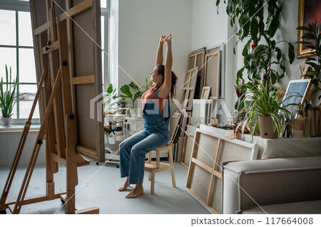 Smiling happy female artist stretching hands up sitting on chair in home art studio workshop. Smiling happy female artist stretching hands up sitting on chair in home art studio workshop. 117664008
