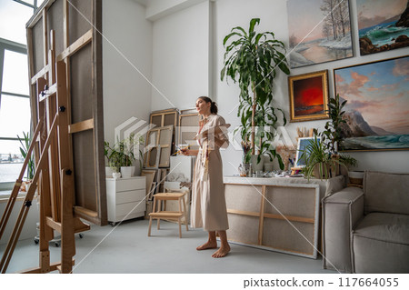 Thoughtful female artist with palette of oil paints and brush stands in front of easel with canvas. 117664055