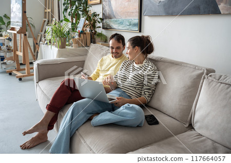 Happy relaxed creative couple of artist smiling sitting with laptop in art studio talking, planning. 117664057
