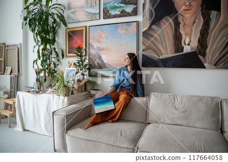 Relaxed woman artist among paintings sitting on sofa couch taking break enjoying tranquillity. 117664058