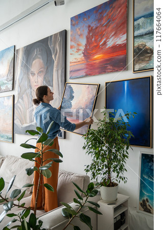 Woman hanging picture on wall. Girl artist painter estimating combination of painting colors. 117664064