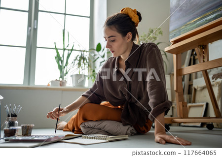 Inspired artistic woman full of ideas embodies fantasies in drawing, sketch painting with aquarelle 117664065