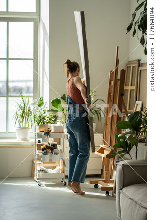 Interested artist young woman holds canvas puts on easel to continue painting in oil in art studio. Interested artist young woman holds canvas puts on easel to continue painting in oil in art studio. 117664094