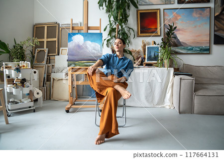 Assured artistic woman, modern contemporary painter taking break from creative work resting on chair Assured artistic woman, modern contemporary painter taking break from creative work resting on chair 117664131