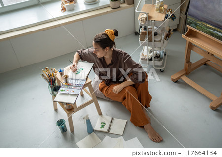Top view of focused woman artist sitting on floor in art studio painting watercolours in sketchbook  117664134