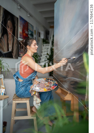Focused modern female artist in home art studio carefully painting, working on huge large artwork. 117664136