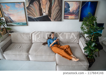 Slothful girl procrastinating lying on sofa with smartphone. Woman relaxing at home art studio. 117664148