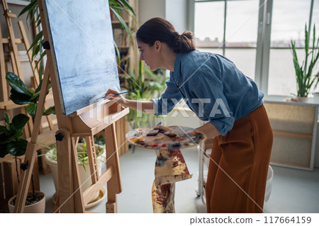 Serious female artist working on artwork masterpiece, deeply immersed in creative art process. Serious female artist working on artwork masterpiece, deeply immersed in creative art process. 117664159