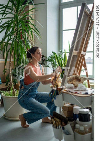 Interested artist female applies oil paint with paintbrush to canvas drawing on easel in art studio. Interested artist female applies oil paint with paintbrush to canvas drawing on easel in art studio. 117664160