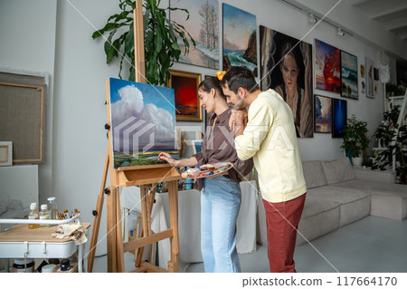 Creative woman artist paints picture in art studio, loving man husband supportive and enjoys process Creative woman artist paints picture in art studio, loving man husband supportive and enjoys process 117664170