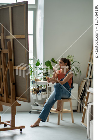 Female artist sits drinking beverage contemplate canvas workshop looking inspiration picture. 117664196