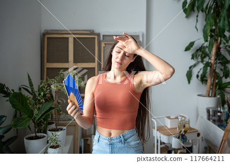 Uncomfortable woman sweats, suffocates, suffers from abnormal weather temperatures, blowing with fan 117664211
