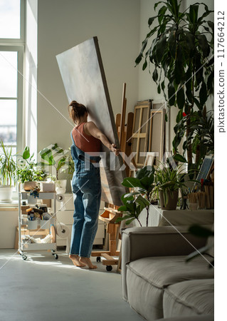 Interested artist young woman holds canvas puts on easel to continue painting in oil in art studio. Interested artist young woman holds canvas puts on easel to continue painting in oil in art studio. 117664212