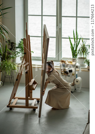 Female artist in bright home art studio organizing preparing workplace, putting workspace in order Female artist in bright home art studio organizing preparing workplace, putting workspace in order 117664213