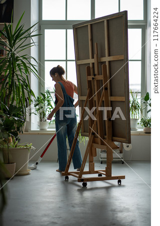 Woman painter carefully cleans in art studio, caring of surroundings at workplace, vacuuming floor. 117664224