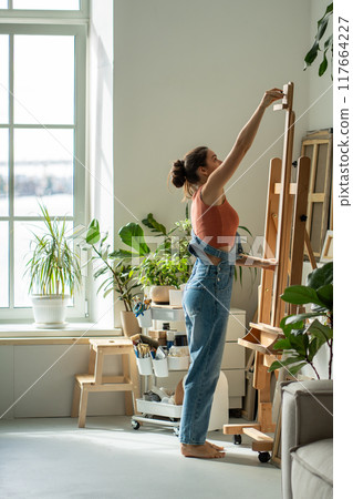 Inspired woman artist preparing easel before work painting process in sunny home studio with plants Inspired woman artist preparing easel before work painting process in sunny home studio with plants 117664227