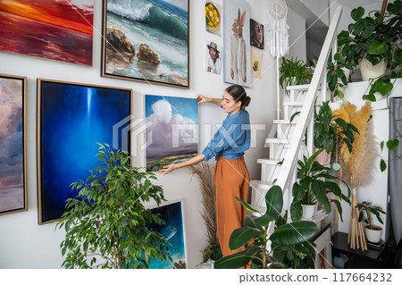 Focused woman artist hanging paintings on wall in creative art home studio workshop apartment. Focused woman artist hanging paintings on wall in creative art home studio workshop apartment. 117664232