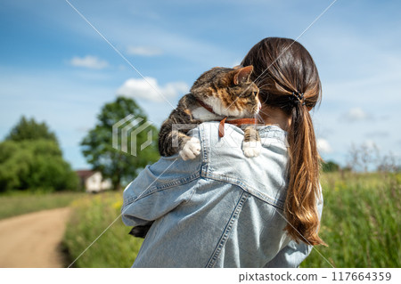 Rear view, woman hugging calm peaceful cat tightly while walking in rural countryside nature area. 117664359