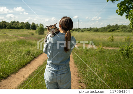 Back view of female owner holding displeased annoyed cat in arms walking among rural green nature. Back view of female owner holding displeased annoyed cat in arms walking among rural green nature. 117664364