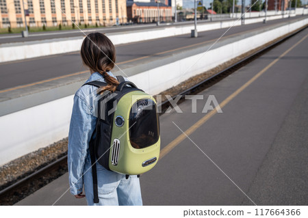 Woman travelling with cat sitting in backpack for pets, standing on platform of railroad station. 117664366