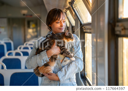 Smiling woman with fluffy tricolor cat traveling riding in electric train together at setting sun. 117664397