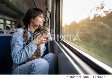 Smiling woman with cat riding on train, looking out of window, enjoying late drive at sunset Smiling woman with cat riding on train, looking out of window, enjoying late drive at sunset 117664400