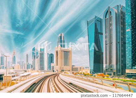 Metro road among glass skyscrapers in Dubai. Metropolitan railway among modern glass skyscrapers in Dubai. Traffic on street in Dubai. Urban background. Altered sky 117664447