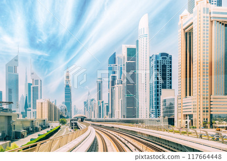Metro road among glass skyscrapers in Dubai. Metropolitan railway among modern glass skyscrapers in Dubai. Traffic on street in Dubai. Urban background. Altered sky 117664448