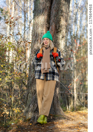 Smiling cheerful female traveler in autumn forest leaning against tree, resting while walking. Smiling cheerful female traveler in autumn forest leaning against tree, resting while walking. 117664468