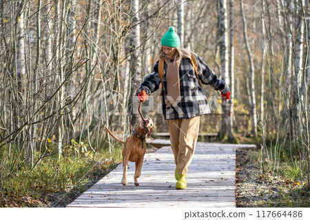 Positive woman walking vizsla dog spends happy weekend in woods eco trail having fun with pet. 117664486