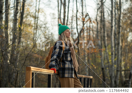 Calm female traveler leaning on railing of bridge in autumn forest, enjoying warm sunny weather. Calm female traveler leaning on railing of bridge in autumn forest, enjoying warm sunny weather. 117664492