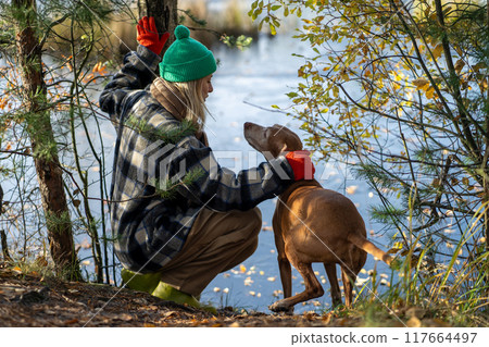 Female traveler with hunting dog sitting on shore by river water, walking in autumn forest together 117664497